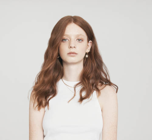 Young woman with long wavy red hair, wearing a white sleeveless top, star earrings, neutral background.
