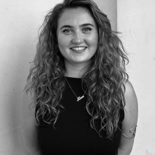 Woman with long curly hair smiling, wearing a sleeveless top and necklace, standing against a plain background.