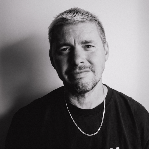 Black and white portrait of a man with a light beard, wearing a chain necklace and dark shirt.