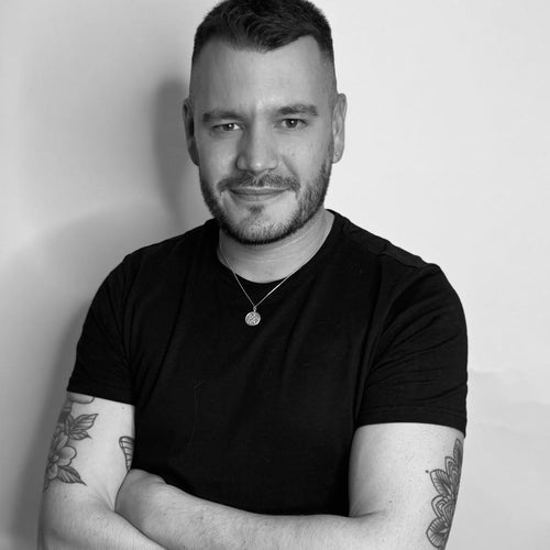 A person with short hair and tattoos, wearing a black shirt, smiles against a plain background.