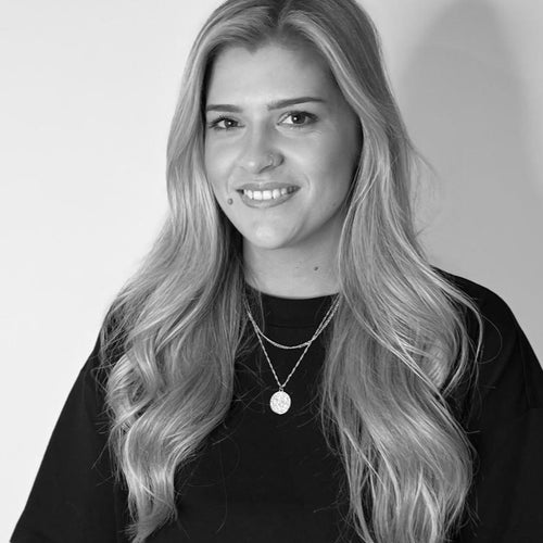Black and white portrait of a smiling woman with long blonde hair, wearing layered necklaces.