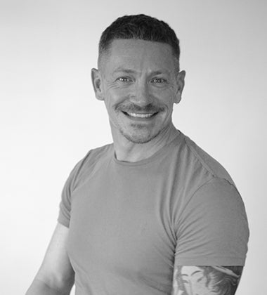 Smiling man with short hair and tattoo, wearing a plain t-shirt, posing in a studio setting.