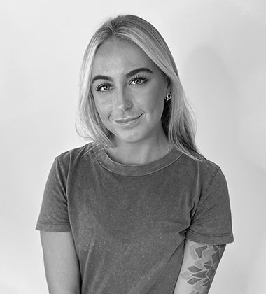 Smiling woman with straight blonde hair, wearing a short-sleeved shirt, showing arm tattoo. Black and white photo.