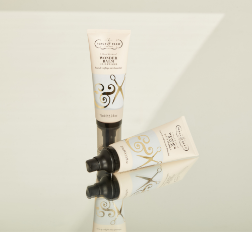 Two tubes of Percy & Reed Wonder Balm, with decorative gold lettering, reflected on a glossy surface.