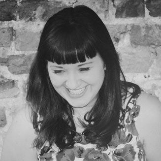 Smiling woman with dark hair and bangs wearing a floral top, against a brick wall background.