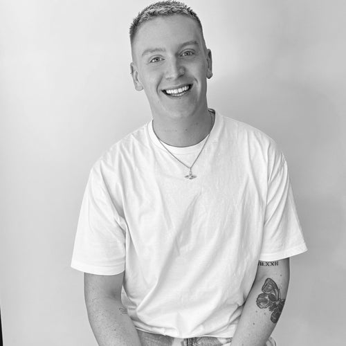 Smiling person with short hair wearing a white t-shirt, necklace, and showing arm tattoos against plain background.